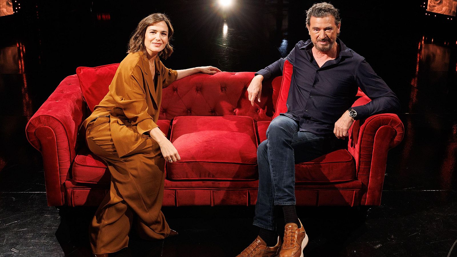 Elena S. Sánchez y Julio Medem sentados en un sofá rojo, ambos sonríen a la cámara en un estudio de televisión.