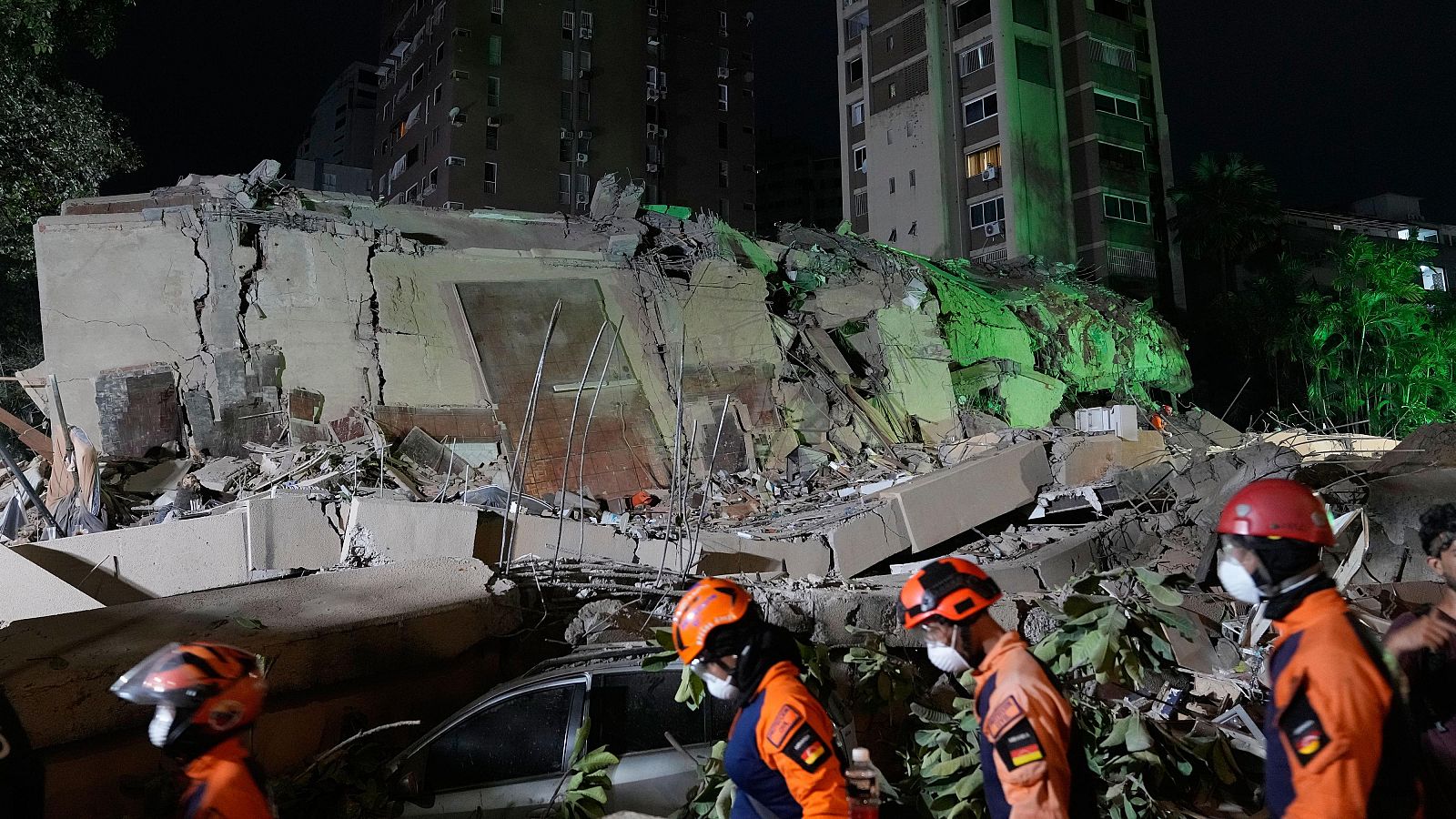 Equips de rescat amb cascos i uniformes lluminosos inspeccionen escombros d'un edifici col·lapsat sota la llum artificial d'una nit fosca.