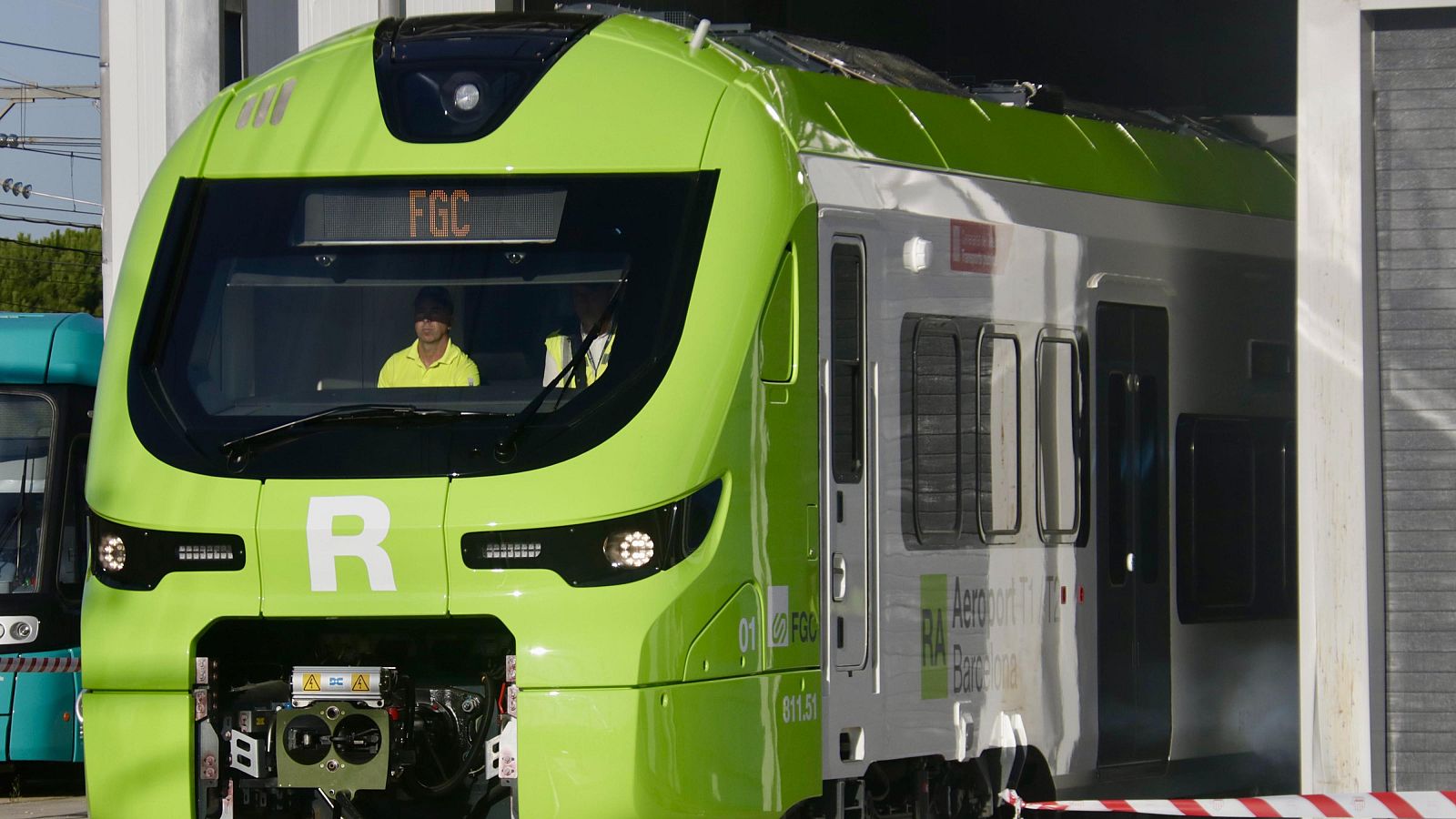 Tren moderno FGC verde y plateado con "RA Aeroport T2 Barcelona" y "01 FGC 611.51". Cabina con conductor de alta visibilidad. Entorno de depósito ferroviario.