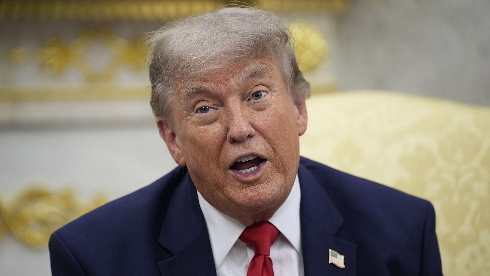 Donald Trump, expresidente de EE.UU., con traje azul, camisa blanca y corbata roja, mira hacia arriba. Lleva un pin de la bandera estadounidense en la solapa. Fondo formal desenfocado.