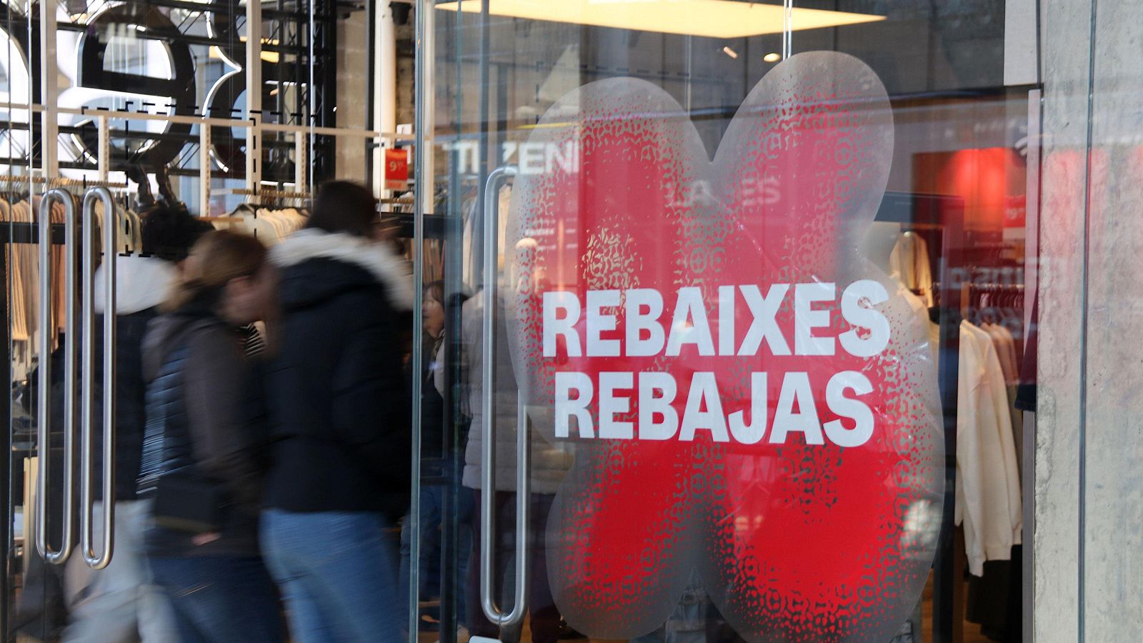 Escaparate de tienda con cartel "REBAIXES REBAJAS" en rojo y blanco. Se aprecian percheros con ropa y personas pasando por delante.