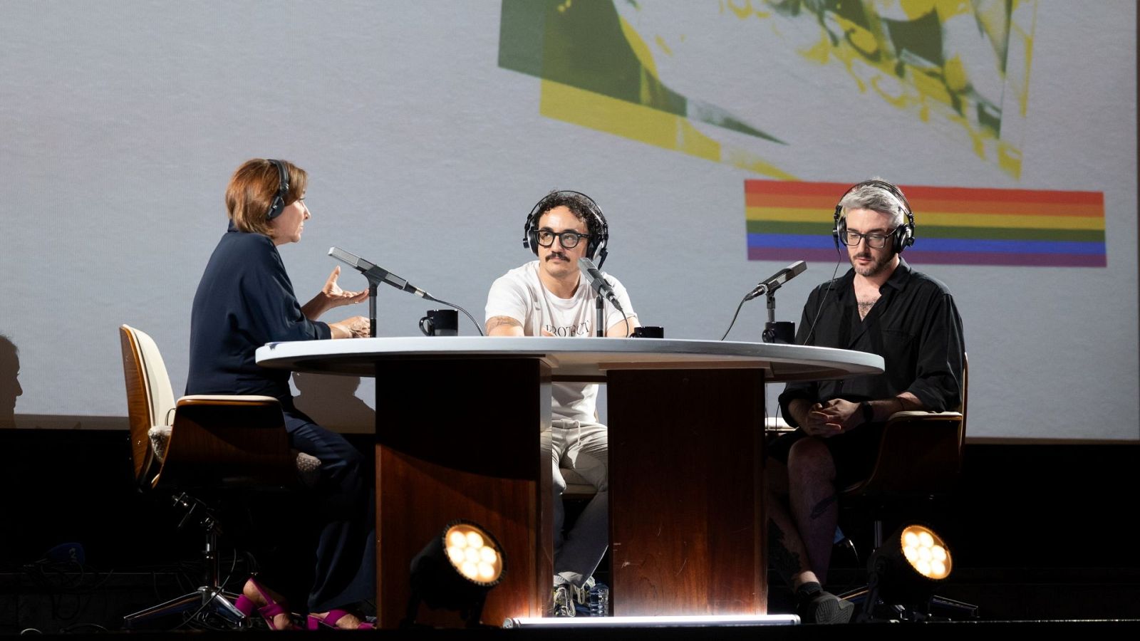 Panel de discusión con Pepa Bueno, César Vallejo y Víctor Mora en un estudio. Pepa gesticula, César y Víctor escuchan atentamente. Fondo con imagen abstracta y bandera LGTBIQ+.