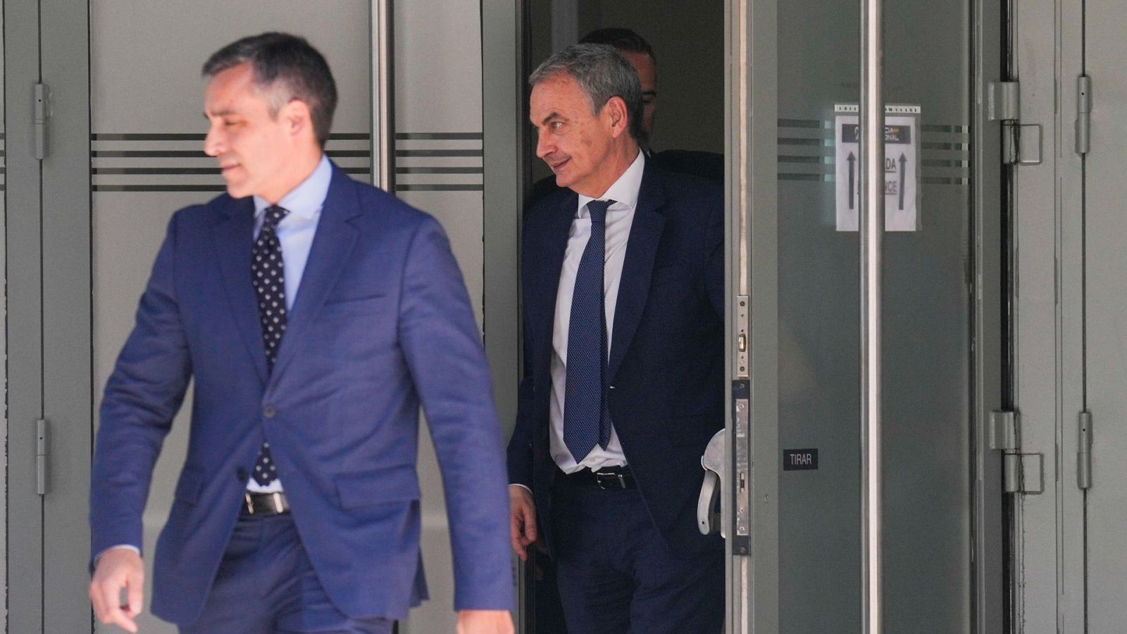 José Luis Rodríguez Zapatero, vestido con traje azul marino, sale de una puerta de cristal con un cartel que dice "TIRAR". Otro hombre camina a su lado.