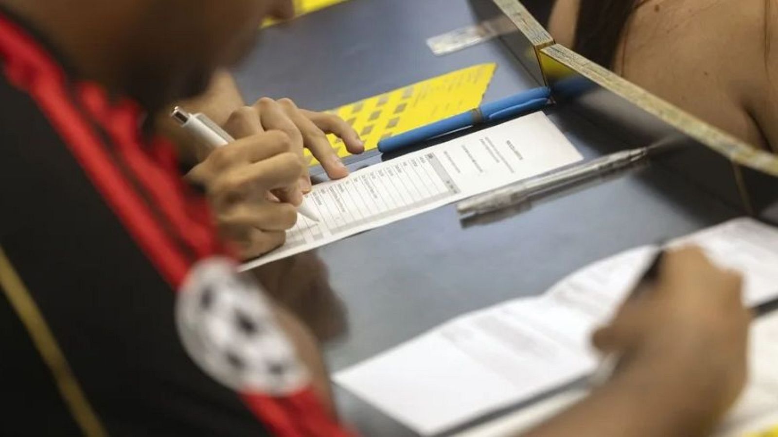 Manos d'un estudiant omplint un formulari de les PAU 2026 amb un bolígraf blanc, sobre una taula fosca amb bolígrafs addicionals i una carpeta groga texturitzada.