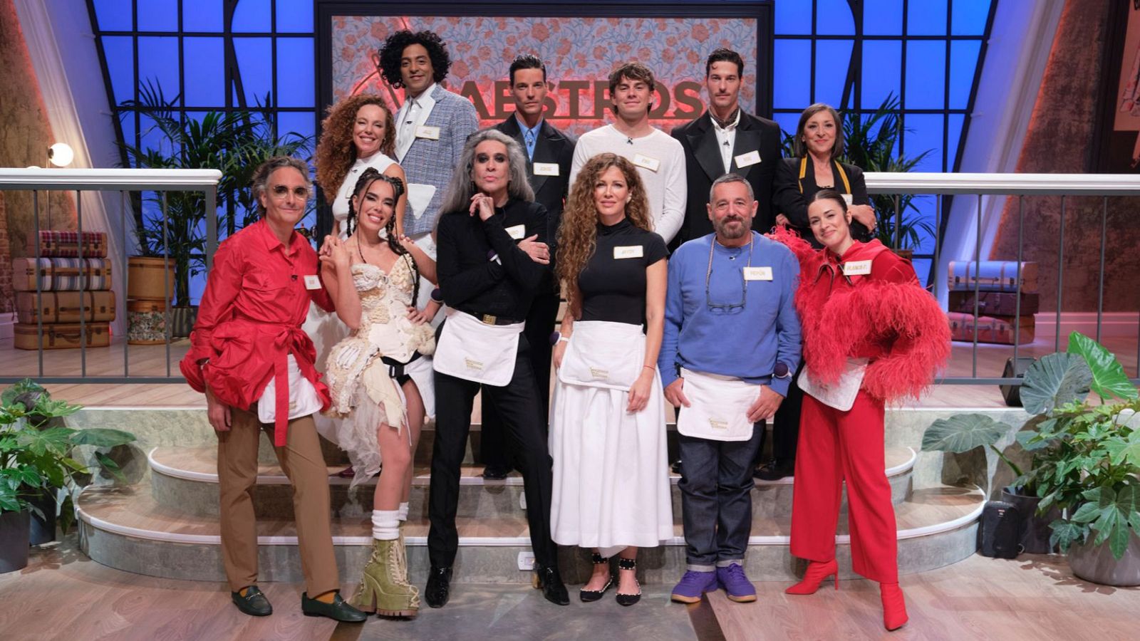 Fotografía grupal de 12 personas, jueces y participantes de "Maestros de la Costura Celebrity 2", en un plató de televisión con decoración de taller de costura. Destacan el logo "MAESTROS" y elementos como plantas y maletas.