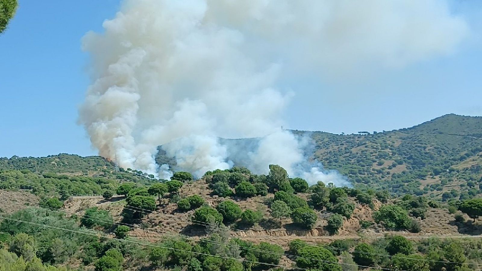 Paisaje natural con colinas verdes y tierra seca, dominado por una columna de humo blanco grisáceo con base rojiza de un incendio activo.