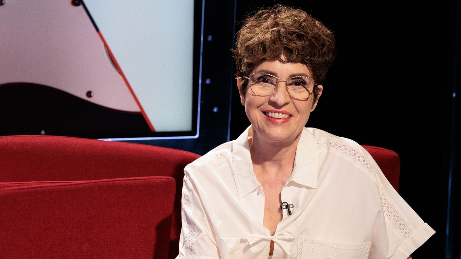 Adriana Ozores, actriz española, sonríe a cámara sentada en un sofá rojo, con camisa blanca, gafas y micrófono. Fondo de estudio de TV con pantalla gráfica.