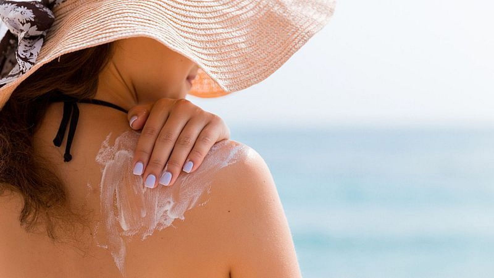 Persona aplicando crema solar en el hombro con uñas lilas, vistiendo sombrero de paja con cinta floral. Fondo de playa soleado.