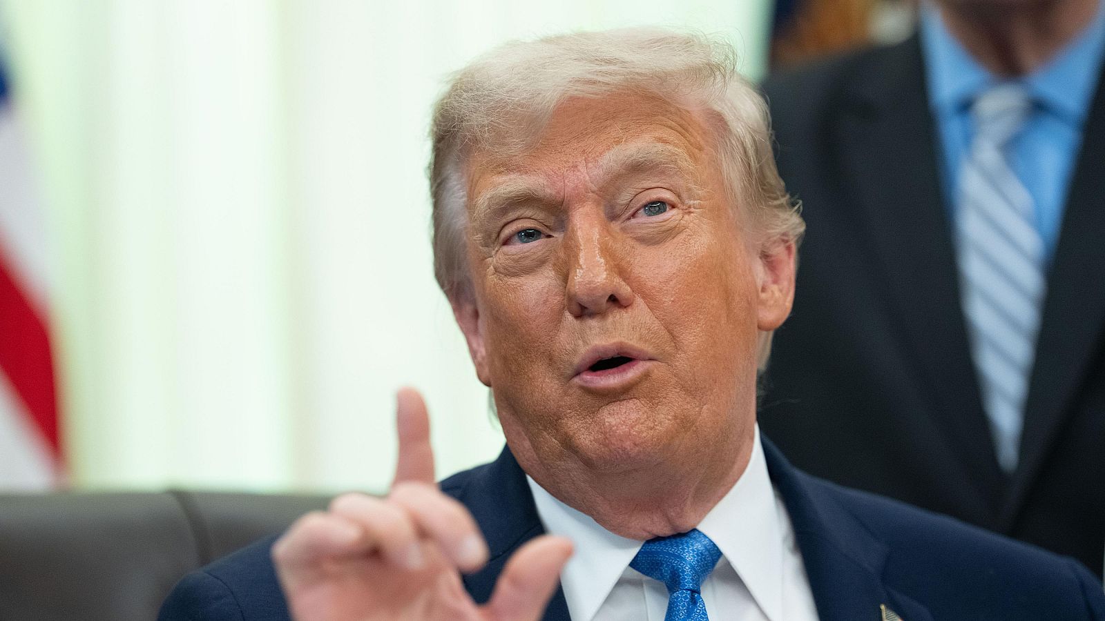 Donald Trump, con traje oscuro y corbata azul, habla con el dedo índice levantado, con la bandera de EE.UU. al fondo.