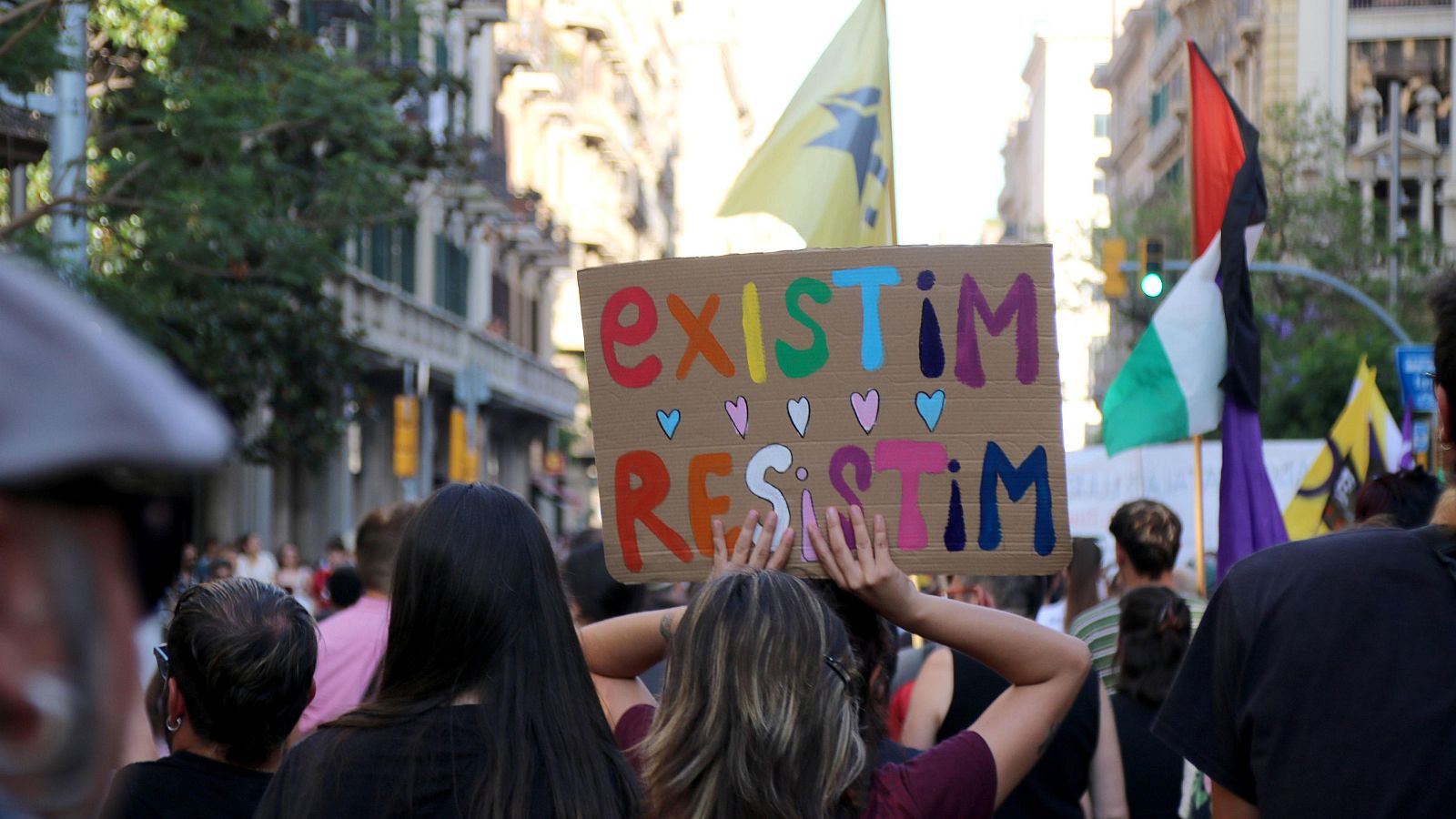 Manifestación urbana con cartel "existIM RESISTIM" y corazones. Se ven la Estelada groga, bandera de Palestina y bandera morada. Calle con edificios, árboles y multitud.