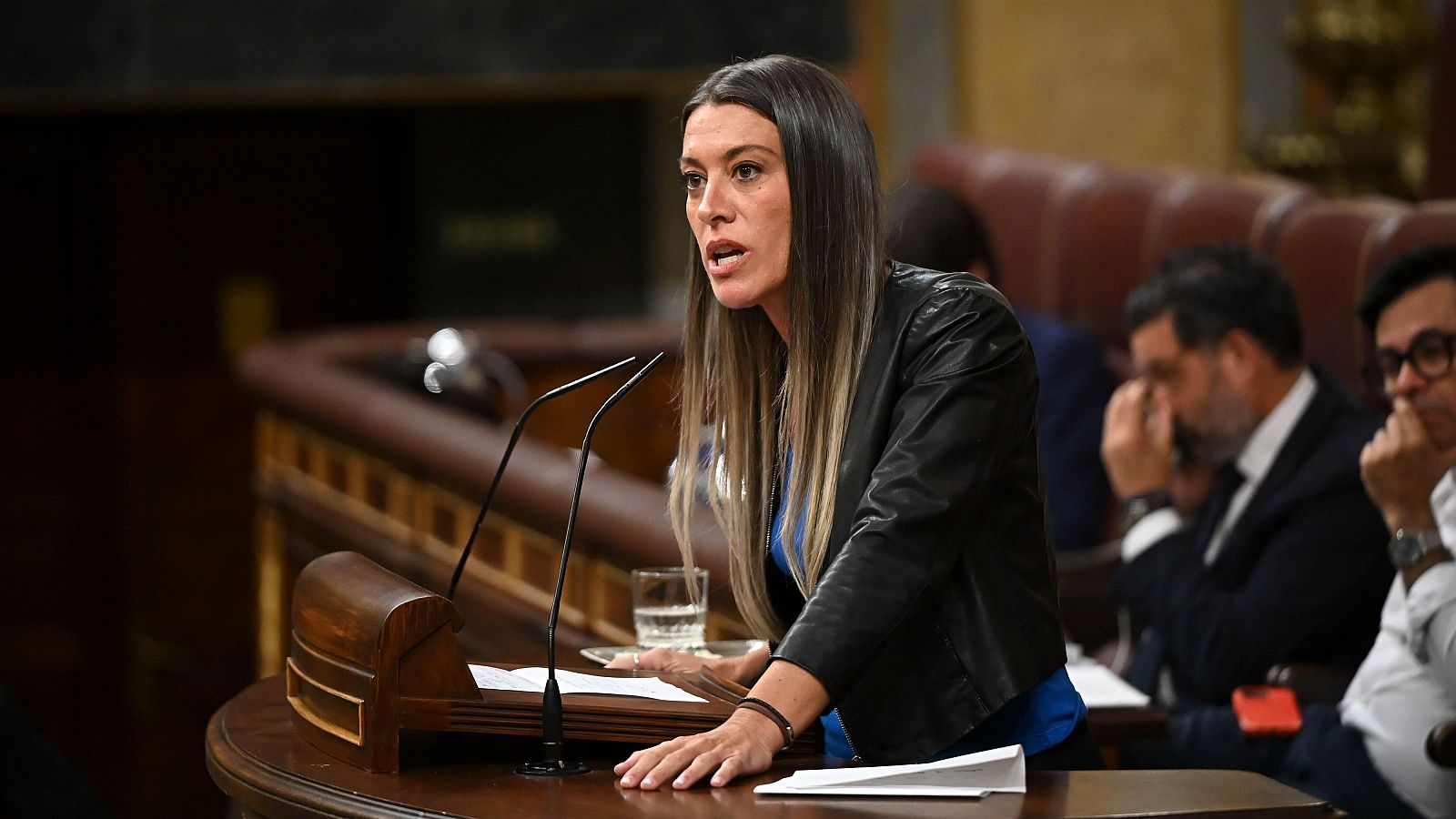 Macarena Olona habla con seriedad detrás de un podio de madera oscura, con micrófonos y folios. Se aprecian dos hombres sentados en segundo plano.