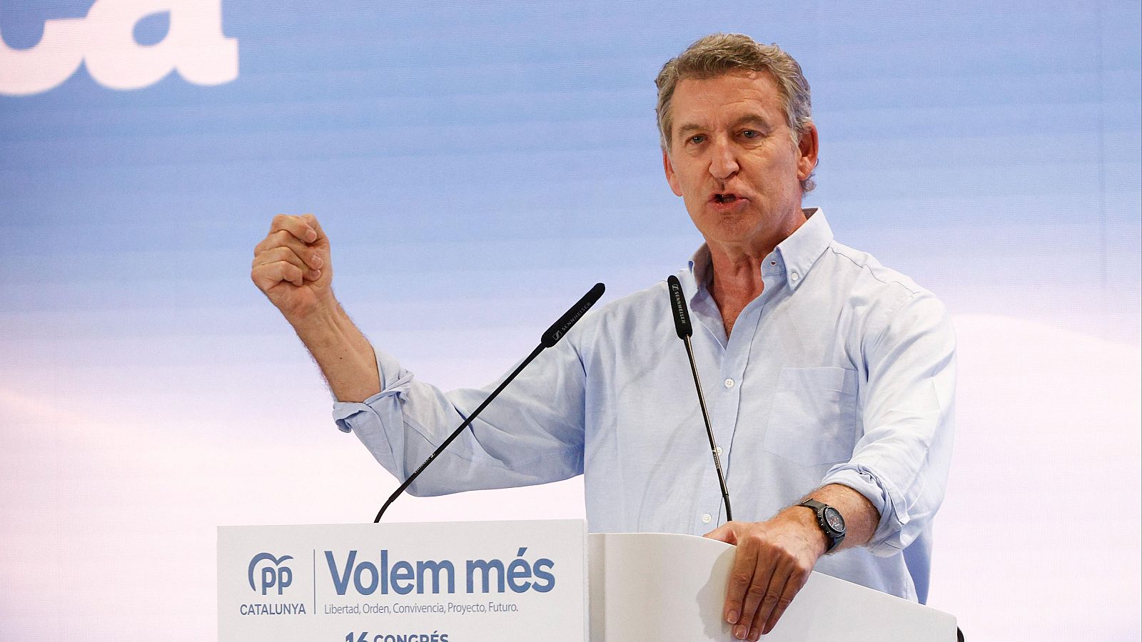 Alberto Núñez Feijóo, líder del PP, habla en el 16º Congreso del PPC en Barcelona. Lleva camisa azul, reloj oscuro y gesticula con el puño cerrado. El podio muestra "Volem més" y principios del partido.