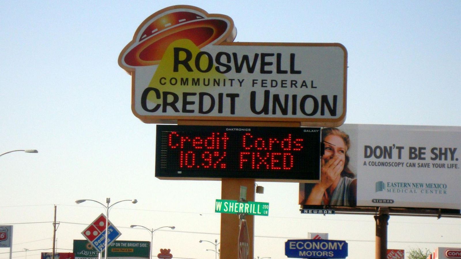 Carteles publicitarios en una calle: "ROSWELL COMMUNITY FEDERAL CREDIT UNION", "Credit Cards 10.9% FIXED", "DON'T BE SHY. A COLONOSCOPY CAN SAVE YOUR LIFE." de "EASTERN NEW MEXICO MEDICAL CENTER", "Domino's Pizza", "ECONOMY MOTORS" y "66" (Ruta 66).