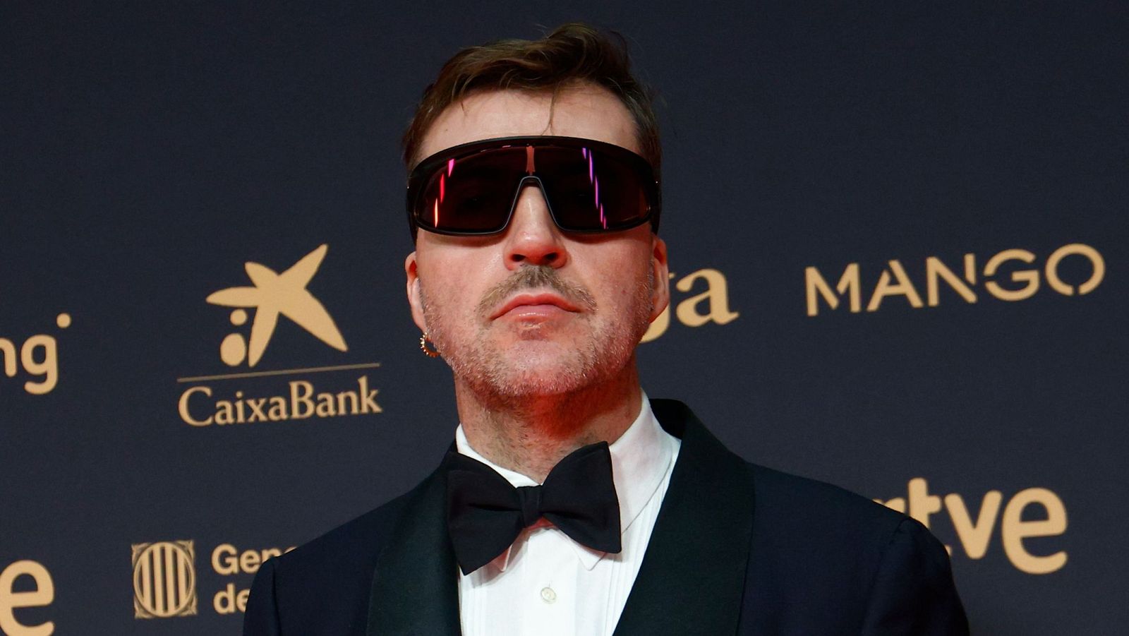 Primer plano de C. Tangana en evento formal con gafas futuristas y esmoquin, rodeado de logos de CaixaBank, MANGO y RTVE.