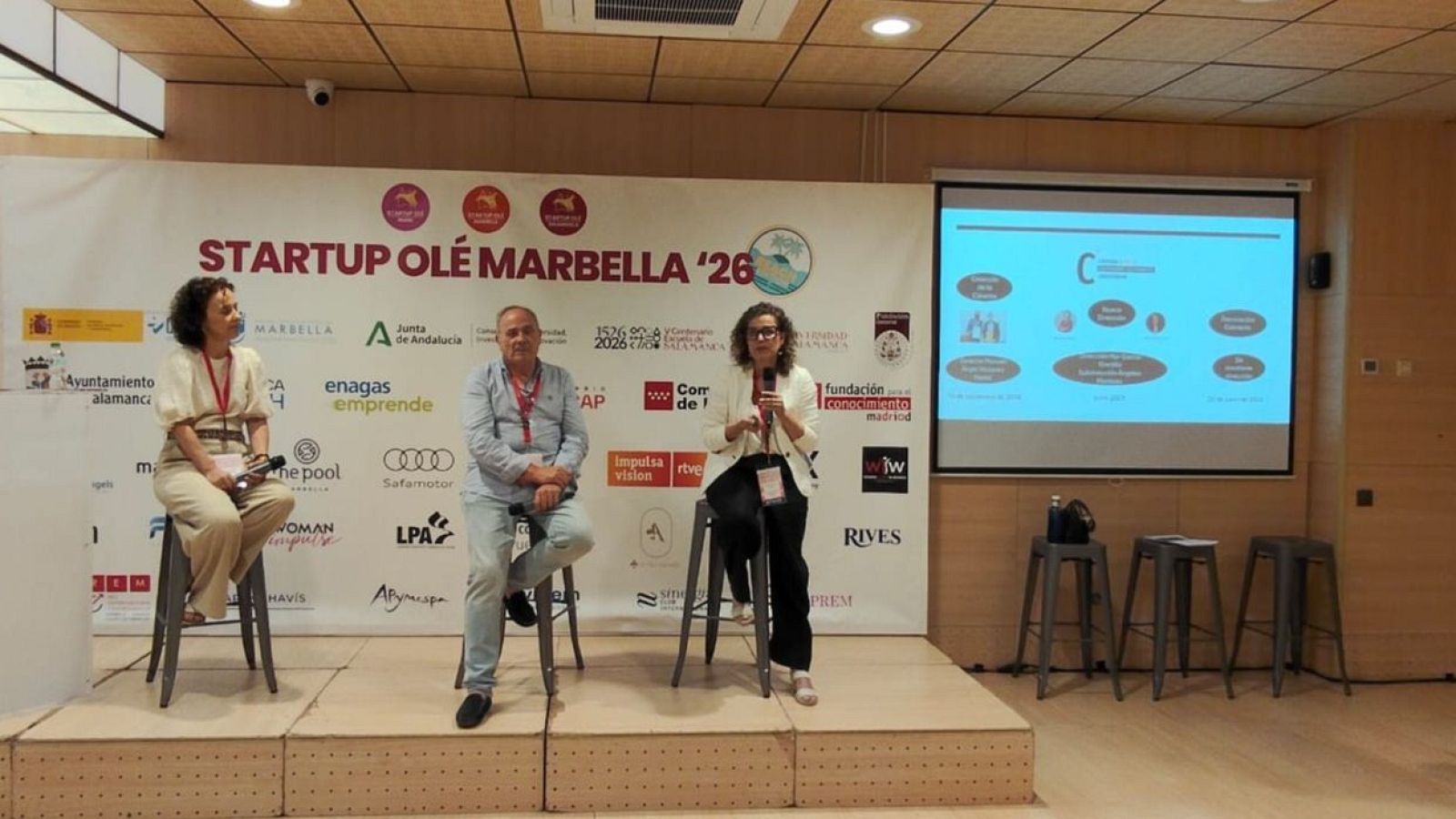 Panel de discusión "MESA DE ESTEBAN" en "STARTUP OLÉ MARBELLA '26" con tres ponentes, un banner de patrocinadores y una pantalla de proyección. Participan "Impulsa Visión".