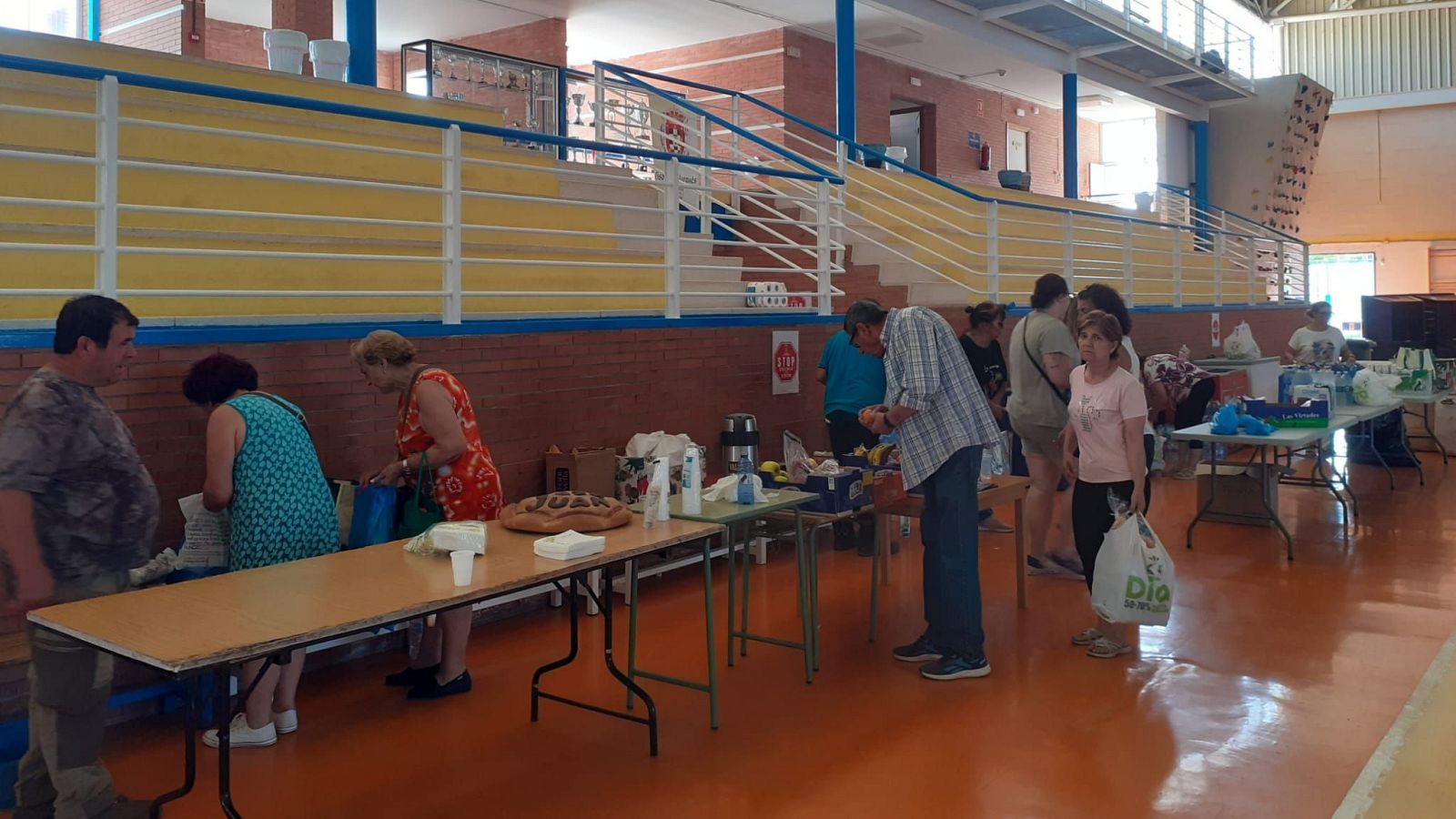 Personas organizan suministros en mesas plegables dentro de un polideportivo con suelo naranja, paredes de ladrillo y una estructura de escaleras con vitrinas.