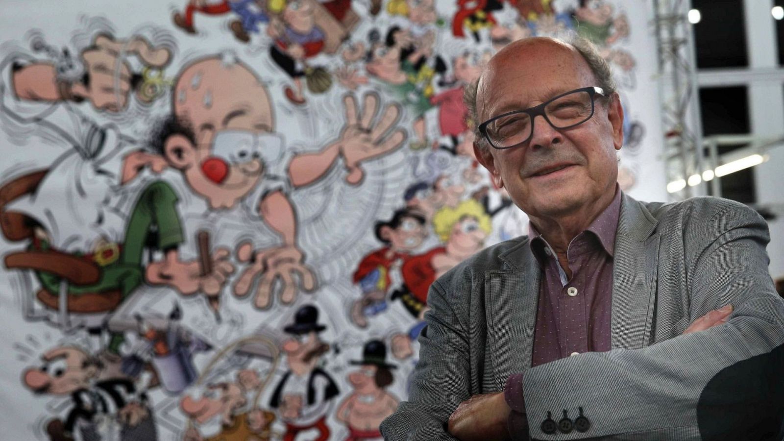 Francisco Ibáñez, creador de Mortadelo y Filemón, sonríe rodeado de sus personajes en un mural. Lleva gafas, camisa granate y chaqueta gris.
