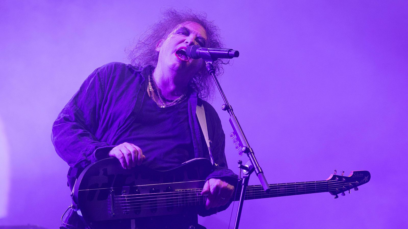 Robert Smith de The Cure, con su distintivo look, canta y toca una guitarra eléctrica oscura bajo una intensa luz púrpura de escenario, con un micrófono frente a él.