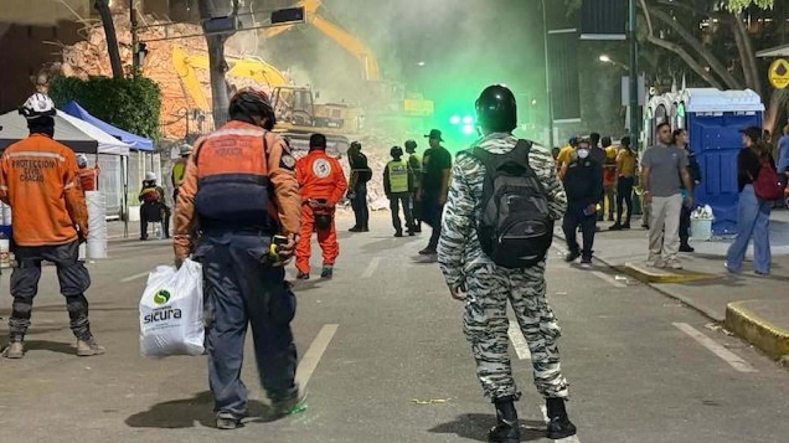 Noche de rescate en Caracas: Protección Civil Chacao, personal con chaleco naranja y militar trabajan entre escombros y excavadoras amarillas, iluminados por luces verdes.