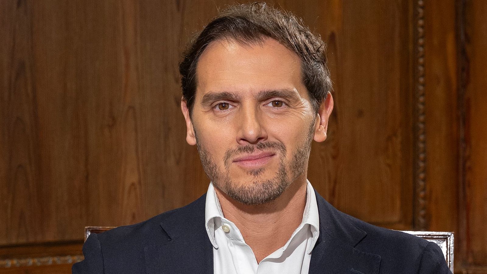 Retrato de Albert Rivera, hombre de mediana edad con barba y bigote, viste camisa blanca y americana oscura, con fondo de paneles de madera.