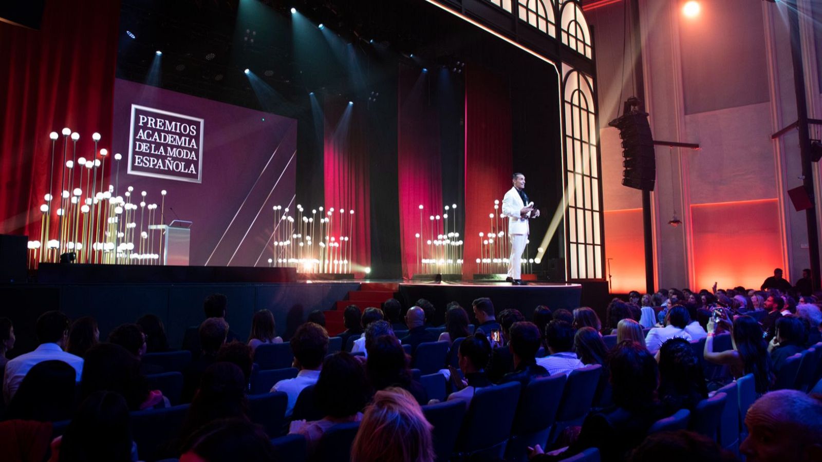 Boris Izaguirre presenta los Premios Academia de la Moda Española en un auditorio, con un escenario iluminado y una audiencia expectante.