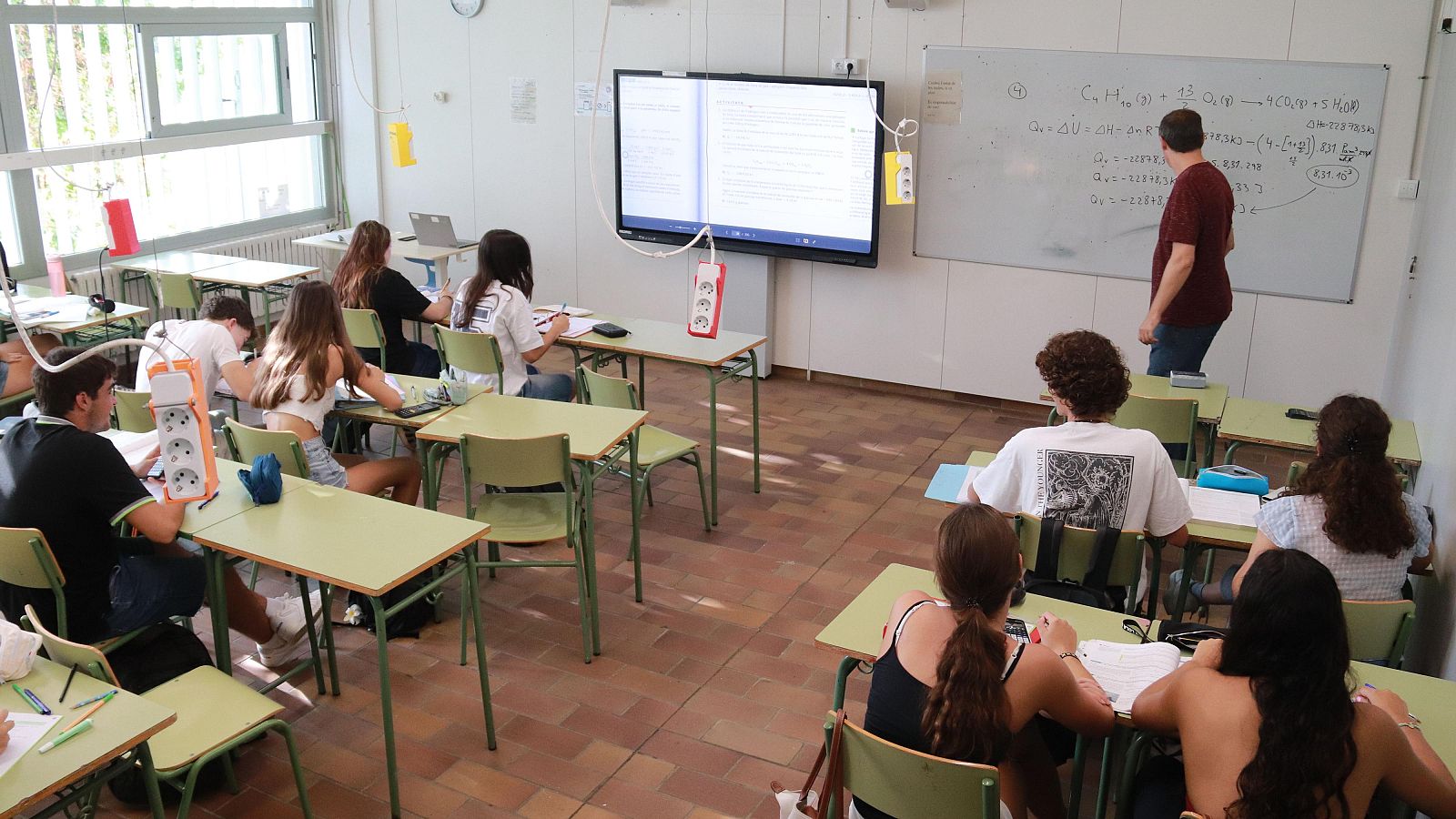 Aula de química con profesor, pizarra con ecuaciones, pantalla interactiva y estudiantes tomando apuntes. Luz natural ilumina el espacio.