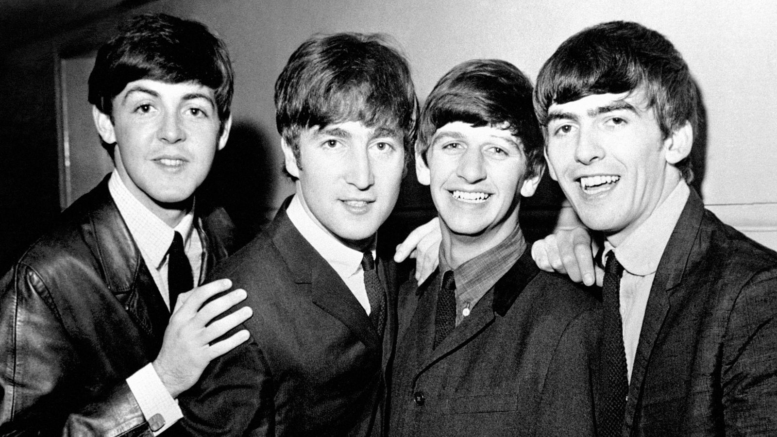Los Beatles (Paul McCartney, John Lennon, Ringo Starr, George Harrison) posan juntos en blanco y negro en 1963, sonriendo y con sus looks icónicos.
