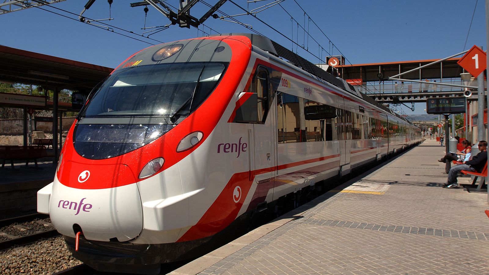 Tren de Renfe blanco y rojo en andén con hombre sentado, banco rojo y señal de andén 1. Puente peatonal naranja al fondo bajo cielo azul.