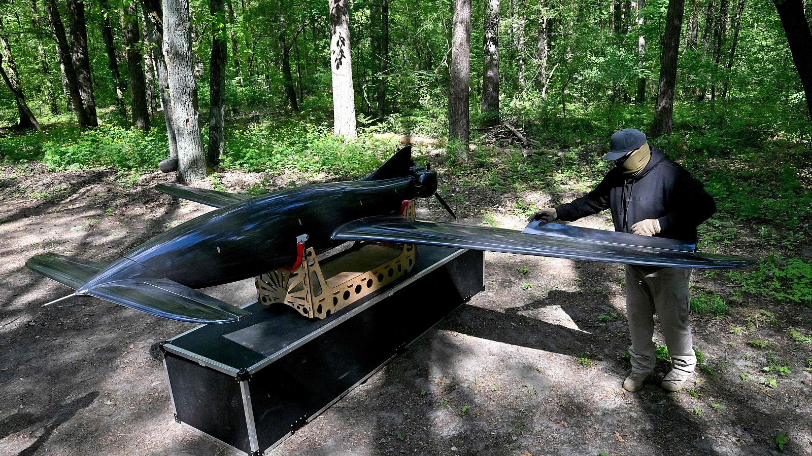 Soldado ucraniano en bosque preparando dron de ala fija oscuro sobre caja de transporte. Rostro cubierto, sudadera oscura, guantes claros.
