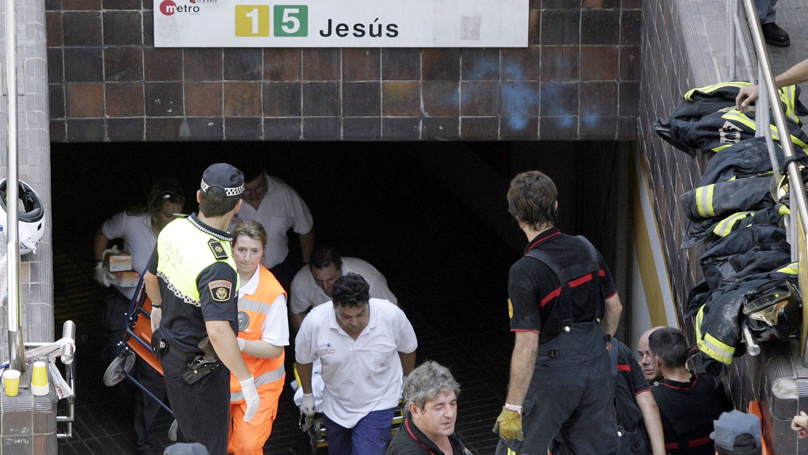 Servicios de emergencia, incluyendo policía, bomberos y personal sanitario, trabajan en la boca de la estación de metro "Jesús" en València, con una camilla de rescate y equipo de bomberos visible.