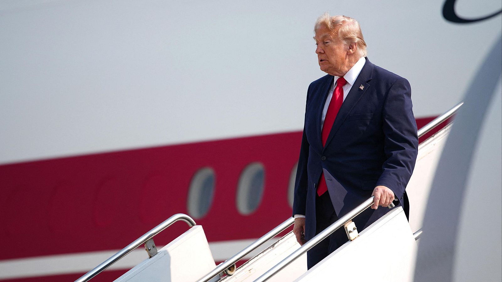 Donald Trump desciende de un avión con traje azul, camisa blanca y corbata roja. El fuselaje del avión es blanco con una franja roja y ventanas.