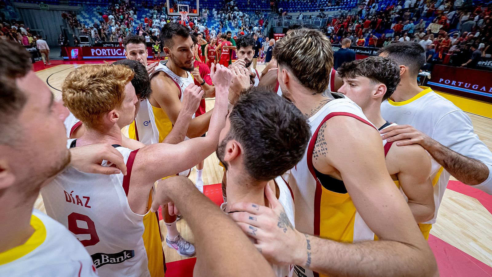 Jugadores de baloncesto abrazados tras un partido contra Portugal. Visten la equipación roja y amarilla. Se ve parte del público y publicidad en la pista.