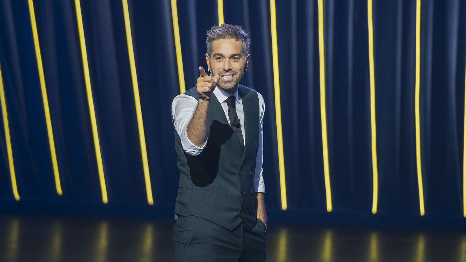 Dani Martínez, presentador, sonríe y señala al frente en un escenario con luces amarillas verticales sobre fondo oscuro. Viste traje oscuro, camisa blanca y corbata.