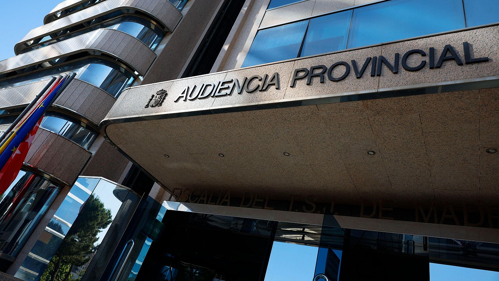 Fachada de la Audiencia Provincial de Madrid con rótulos "AUDIENCIA PROVINCIAL" y "FISCALÍA DEL ESTADO DE MADRID", flanqueada por las banderas de España y la Comunidad de Madrid bajo un cielo azul.