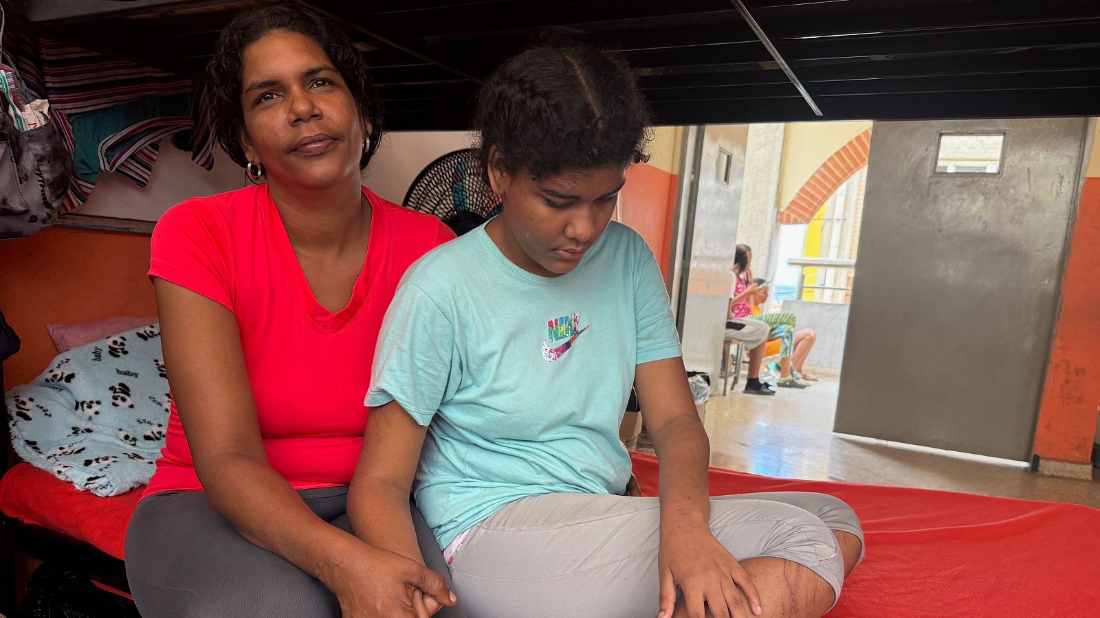 Jennibel y Sofía, madre e hija, sentadas juntas en un centro de acogida. Jennibel viste camiseta roja y Sofía una azul con logo NIKE.