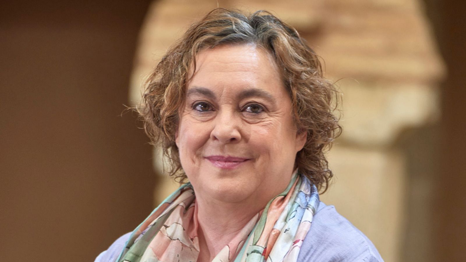 Retrato de Laila Ripoll, dramaturga y directora de teatro, con cabello castaño rizado y bufanda de seda colorida. Fondo desenfocado con tonos cálidos.