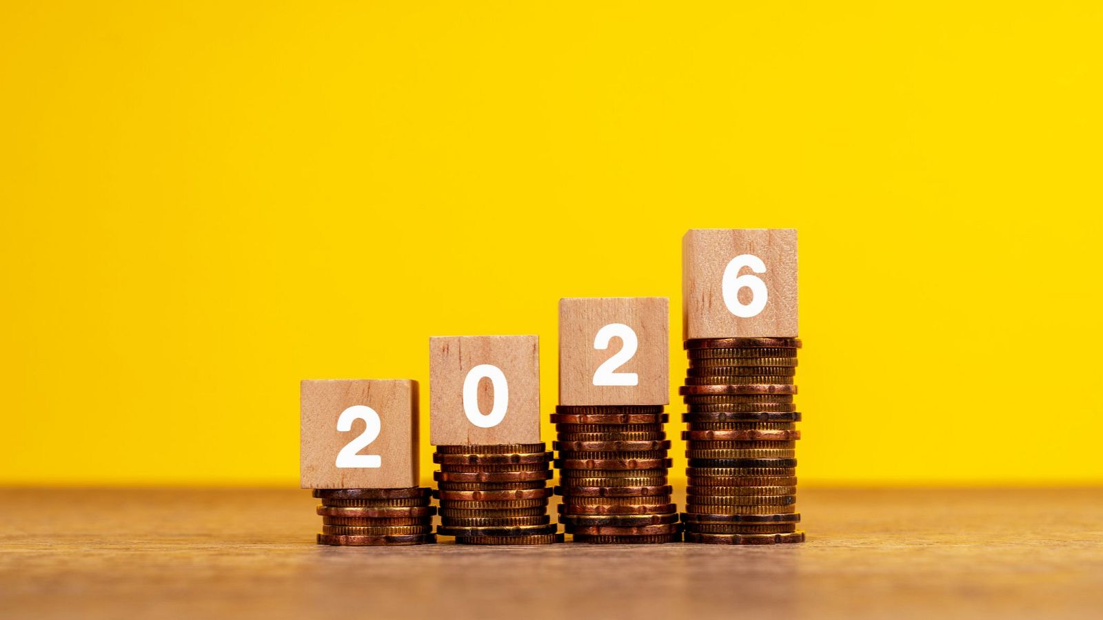 Cuatro pilas de monedas ascendentes coronadas por bloques de madera con los números 2, 0, 2, 6, formando el año 2026, sobre fondo amarillo y base de madera.