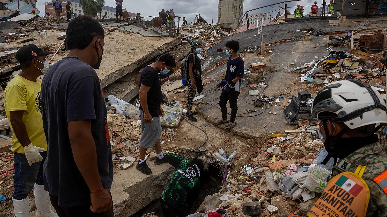 Escena de devastación urbana con rescatistas y voluntarios buscando entre escombros. Se observa un rescatista con distintivos de "AYUDA HUMANITARIA MÉXICO EJÉRCITO FUERZA AÉREA".