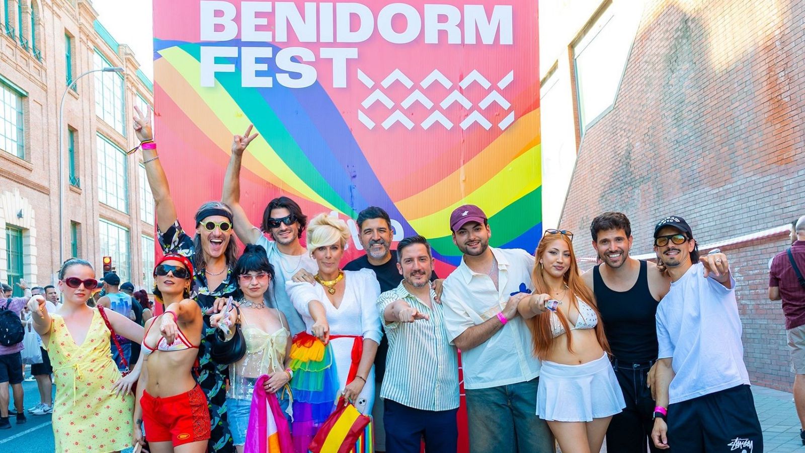 Artistas del Benidorm Fest, junto a María Eizaguirre, Tony Aguilar y Sergio Calderón, celebran el Orgullo frente a un cartel con el logo del festival y los colores del arcoíris.
