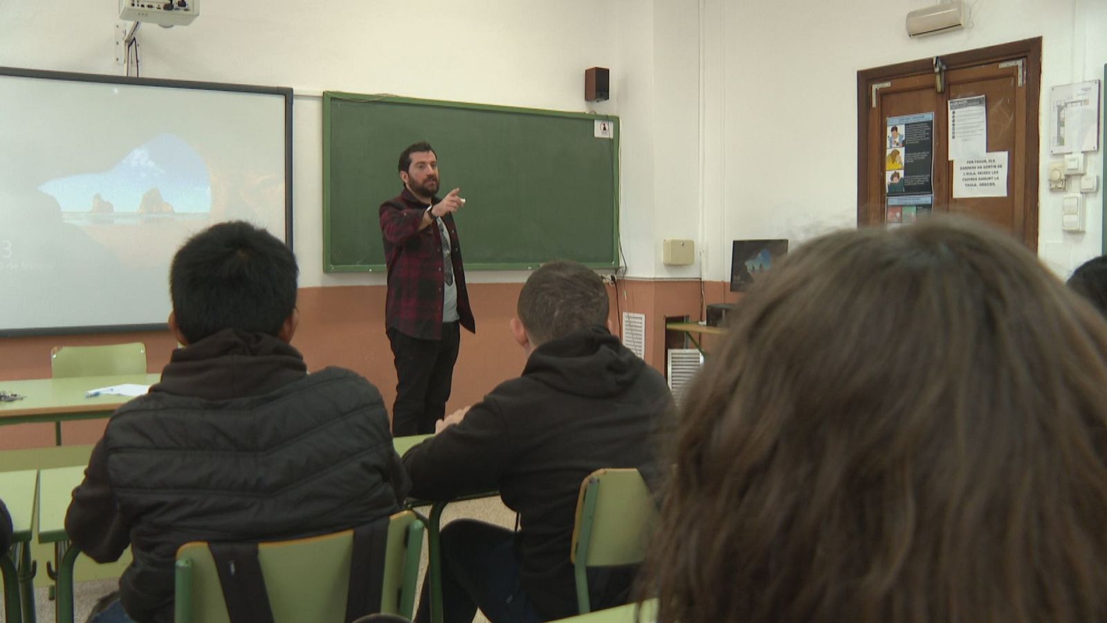 Instructor con barba y camisa a cuadros explica en un aula con pizarra vacía y pantalla de paisaje. Estudiantes sentados de espaldas atienden.
