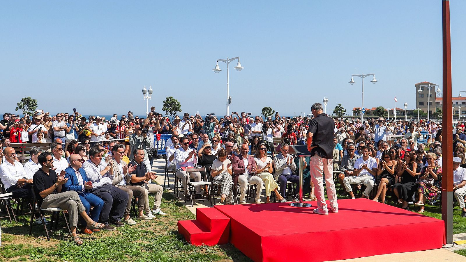 Homenaje a Luis Enrique en Asturias. El homenajeado, de espaldas, se dirige a una multitud entusiasta en un escenario al aire libre, con césped y agua al fondo.