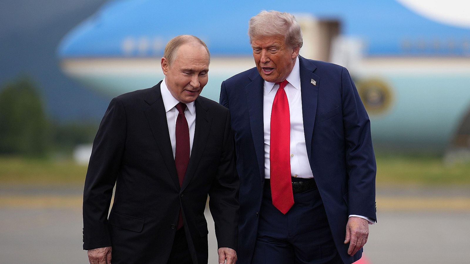 Donald Trump y Vladímir Putin caminan en una base aérea. Trump viste traje azul y corbata roja; Putin, traje oscuro y corbata burdeos. Un avión y montañas al fondo.