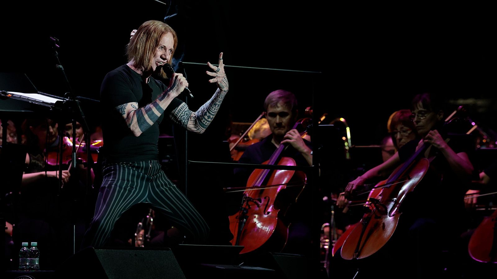 Danny Elfman, con cabello rojizo y tatuajes, canta en un micrófono en un escenario oscuro, acompañado por la Orquesta de RTVE.