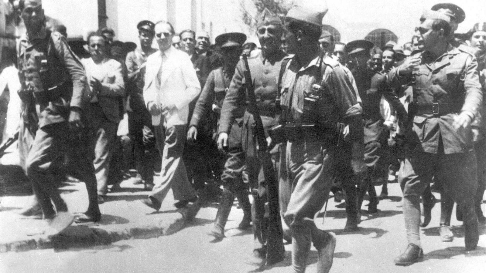 Francisco Franco, con uniforme militar, camina por una calle urbana acompañado de un hombre sonriente y un séquito. La imagen data de la década de 1930.