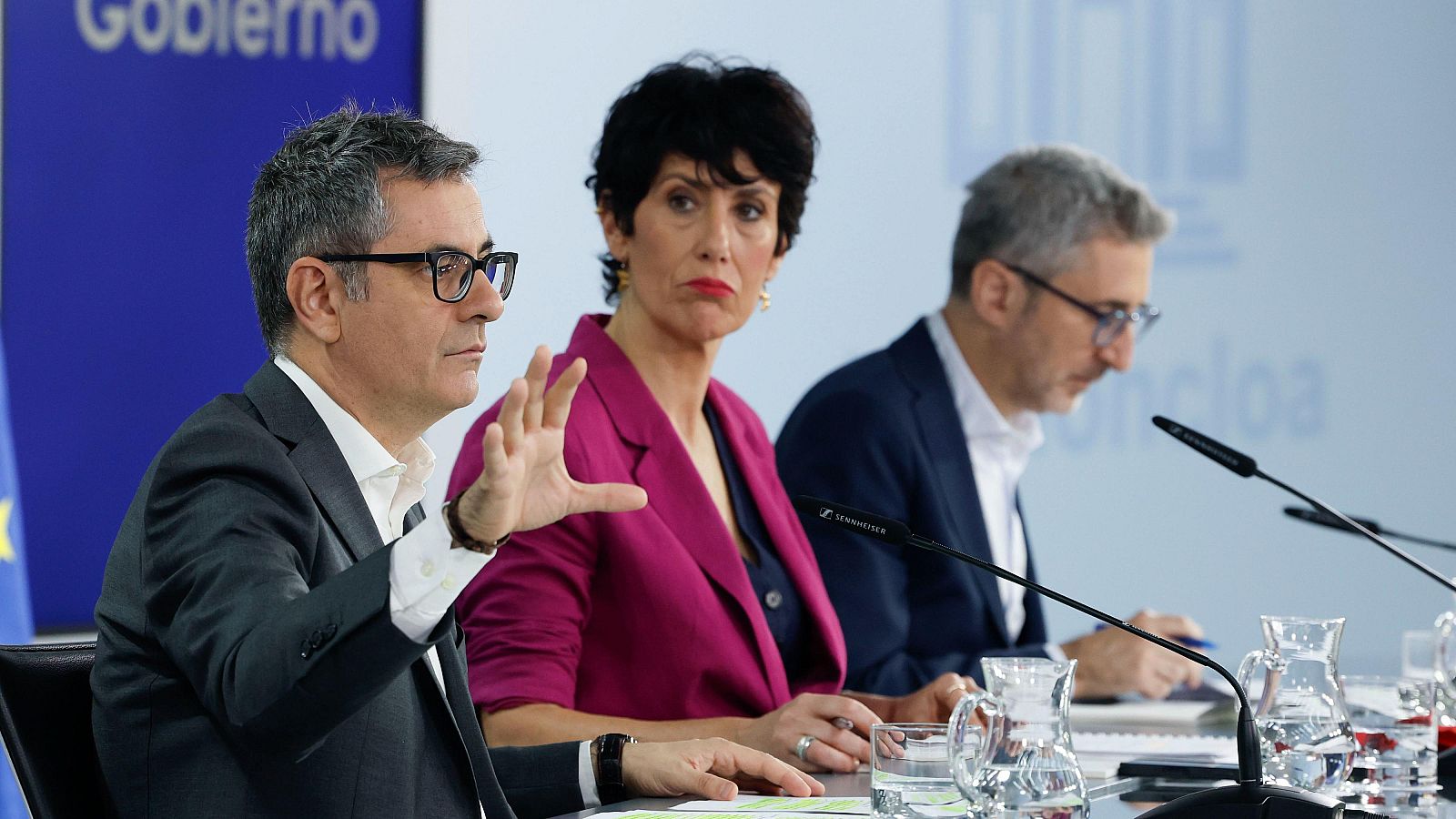 Félix Bolaños, Elma Saiz y Arcadi España en una rueda de prensa. Se aprecian micrófonos, vasos y documentos sobre la mesa.