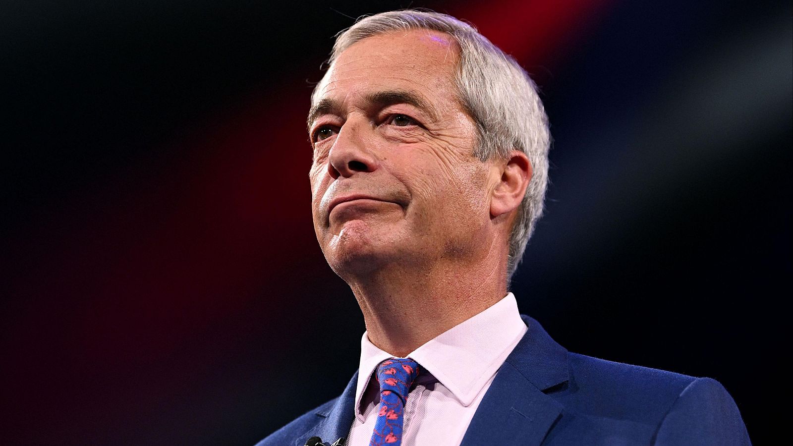 Primer plano de Nigel Farage con traje azul marino, camisa rosa pálido y corbata azul con patrón anaranjado, durante un congreso en septiembre de 2025. Expresión seria, micrófono de solapa visible.