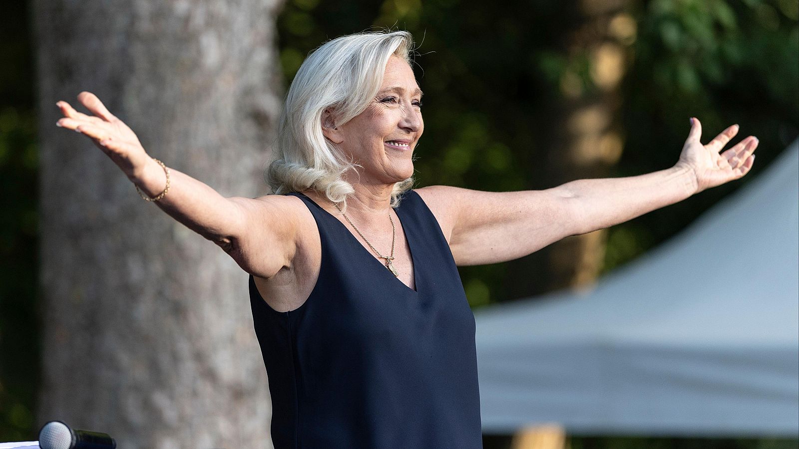 Marine Le Pen saluda a la multitud en un mitin en Liévin, Francia. Sonríe ampliamente con los brazos extendidos, luciendo su cabello rubio platino y un top azul marino con collar dorado. La imagen transmite entusiasmo y conexión.