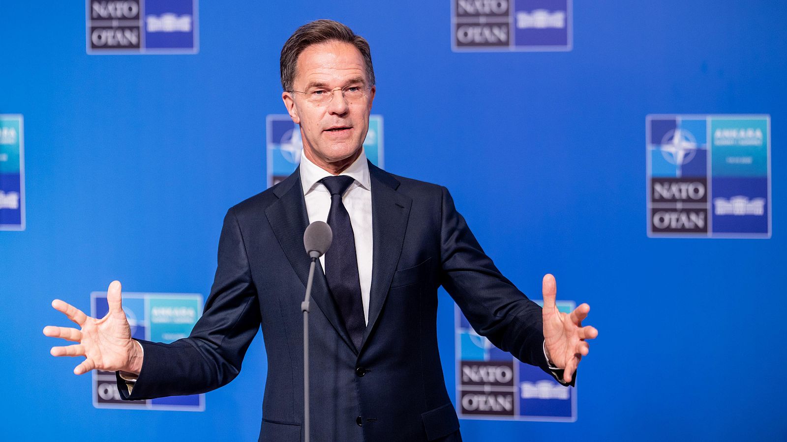 Mark Rutte, secretario general de la OTAN, habla en un evento, gesticulando con las manos. Viste traje oscuro, camisa blanca y corbata azul. Fondo azul con logos "NATO OTAN" y "ANKARA".