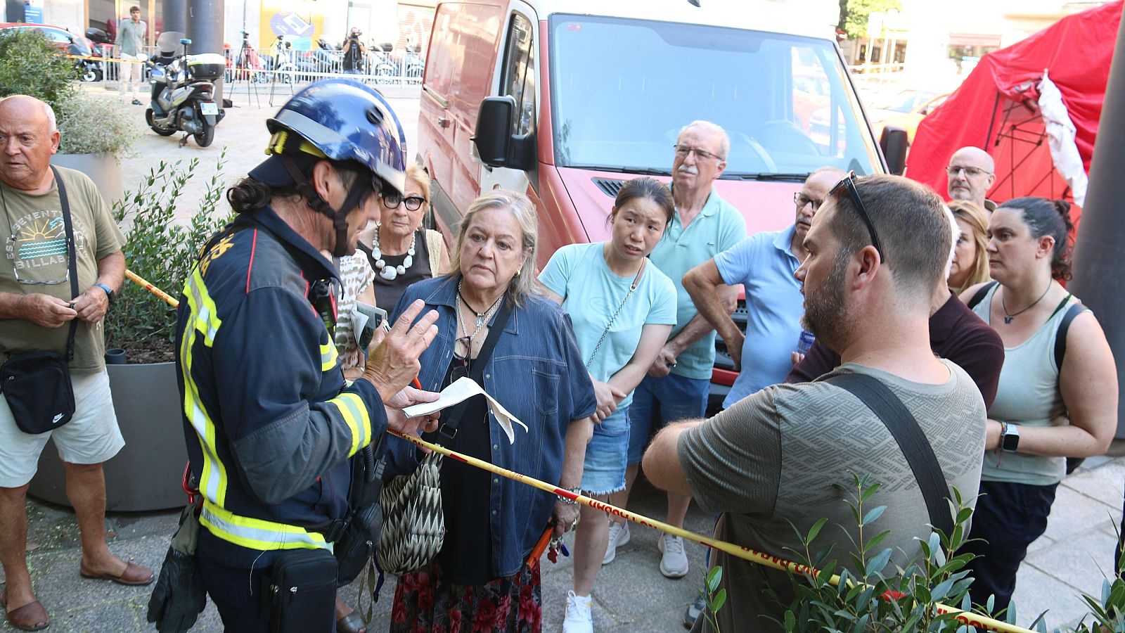 Bombero informa a civiles en calle urbana con vegetación, furgoneta y carpa roja. Cinta de balizamiento indica zona restringida.