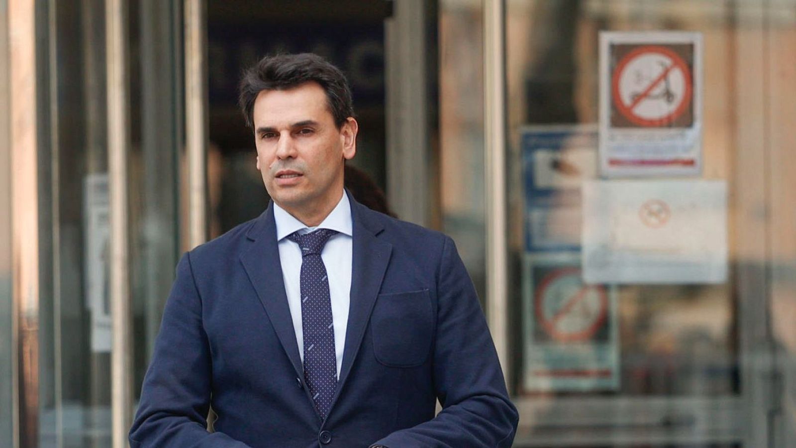 Hombre con traje azul marino, camisa azul claro y corbata oscura con lunares blancos sale de un edificio con puertas de cristal y señales, entre ellas una de prohibición de paso con patinete.